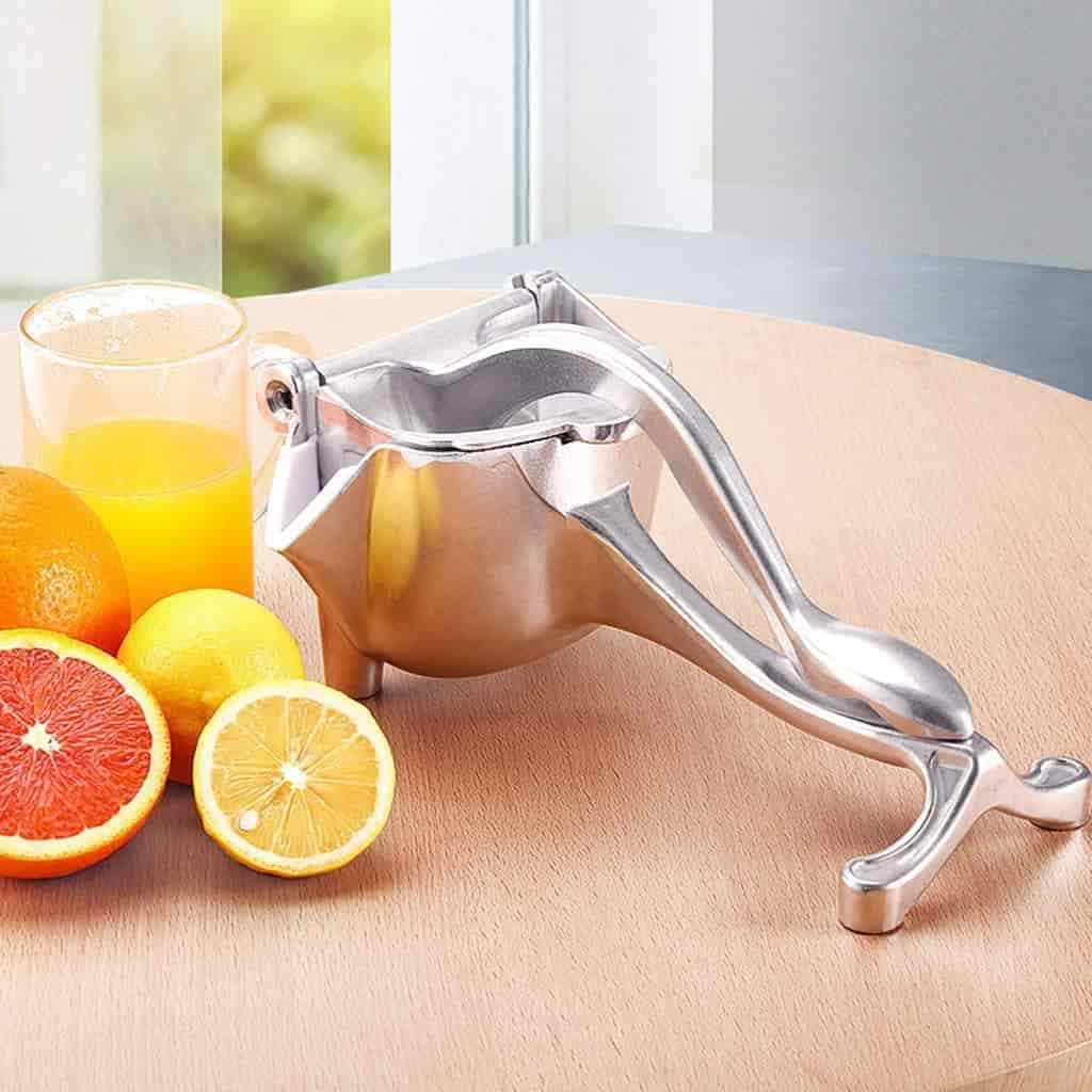 Metallic lemon squeezer on a wooden surface with lemons and a glass of juice in the background.