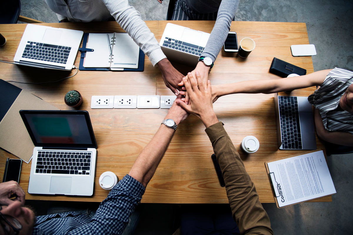 Overhead view of several people stacking their hands together on a wooden conference table, surrounded by laptops, coffee cups, and office documents.